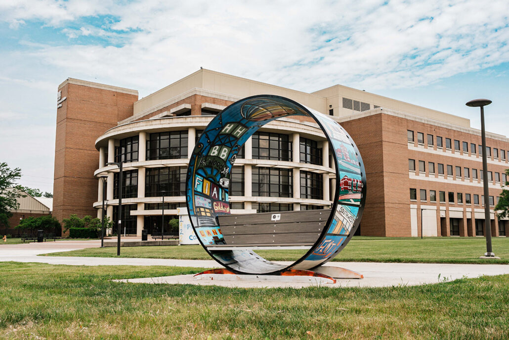 A colorful circular bench sculpture in front of a modern building, surrounded by grass and trees.