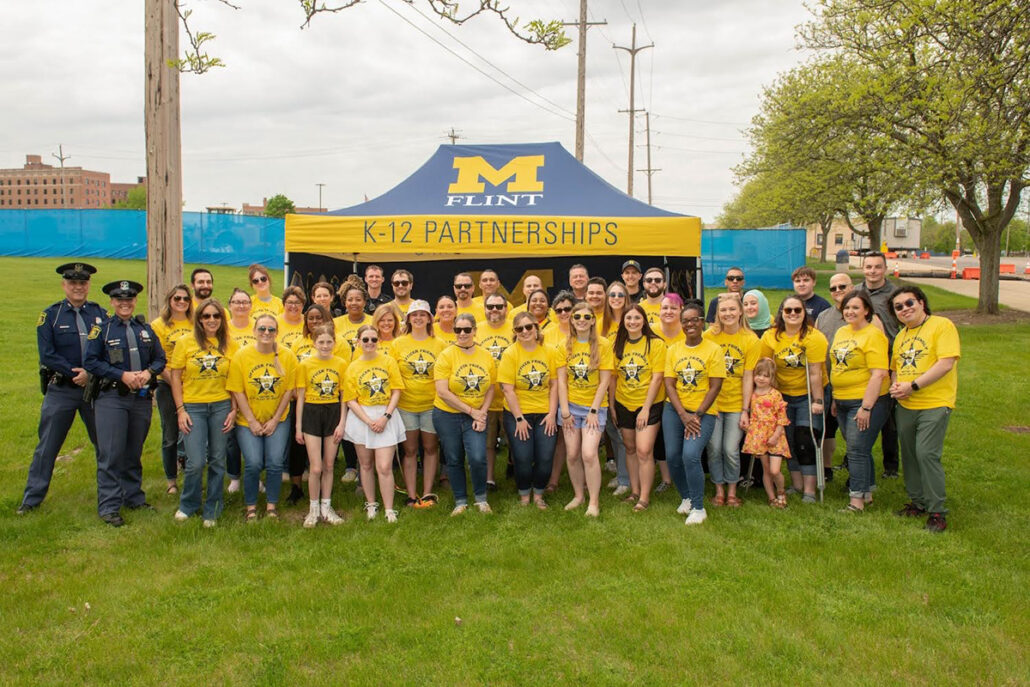 A group of people wearing yellow shirts with a star design stands in front of a tent that reads "K-12 PARTNERSHIPS" and "M FLINT."