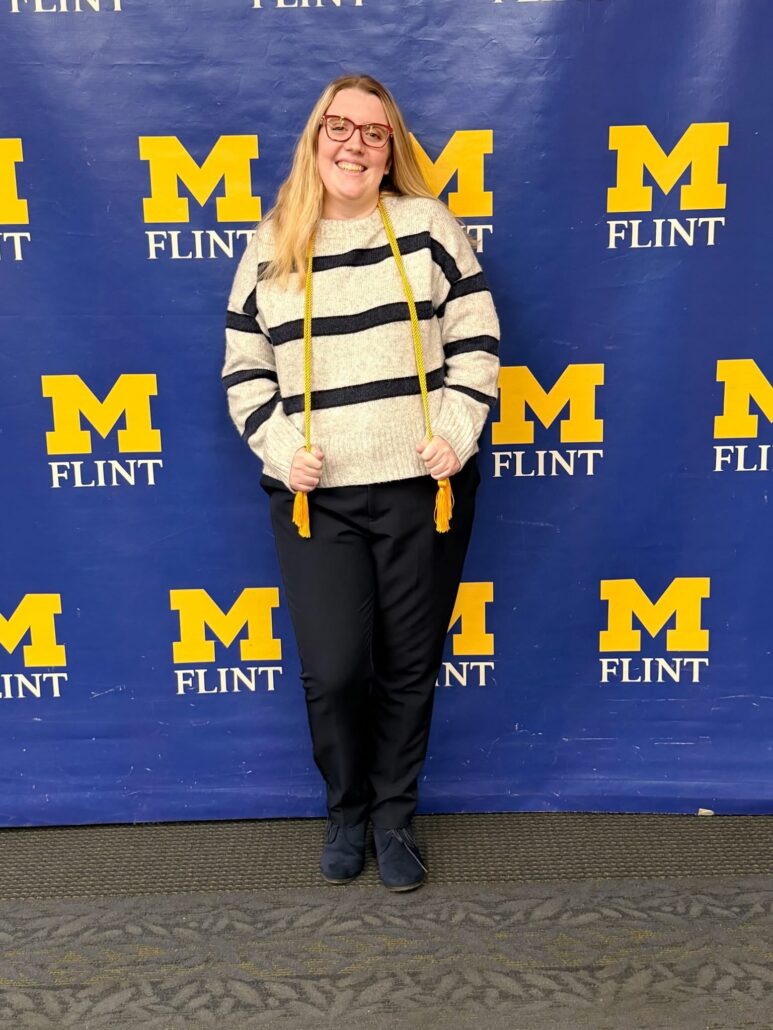 A person stands in front of a blue backdrop with yellow "M FLINT" lettering, wearing a striped sweater and holding a yellow cord.