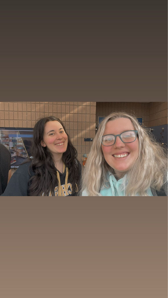 Two individuals are posing for a selfie, one with long dark hair wearing a black hoodie, and the other with wavy blonde hair in a light blue hoodie. The background features a wall with a display.