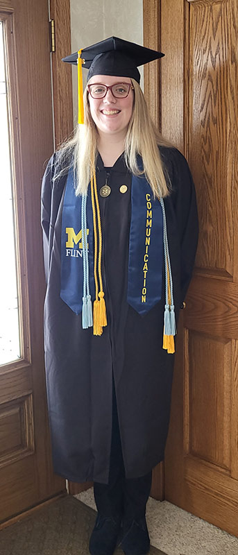A person wearing a black graduation gown and cap, with blue and yellow stoles that read "M" and "COMMUNICATION," standing in front of a wooden door.