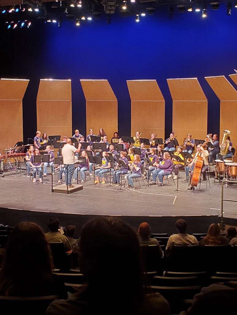 A conductor leads UM-Flint's Wind Symphony in a performance on an auditorium's stage