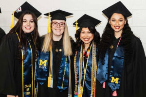 Four graduates in black caps and gowns pose together, wearing blue and gold stoles. One stole reads "CLASS OF 2025" and another features "COMMUNICATION."