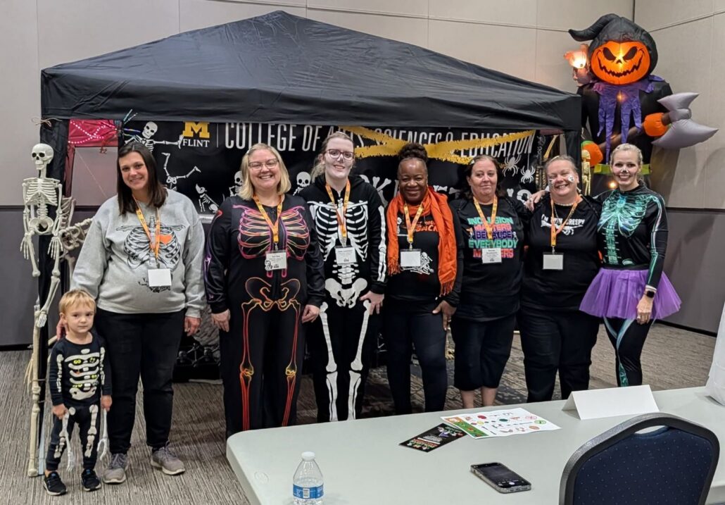 A group of people dressed in Halloween-themed outfits, including skeleton costumes, stands in front of a decorated booth with a black canopy. A large inflatable pumpkin and skeletons are visible in the background. A table with a water bottle and flyers is in the foreground.