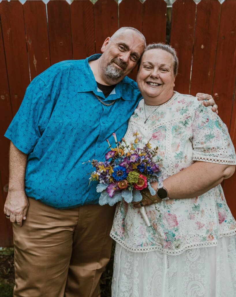A person in a blue patterned shirt stands next to another person in a floral dress, holding a bouquet of colorful flowers. They are posing in front of a wooden fence.