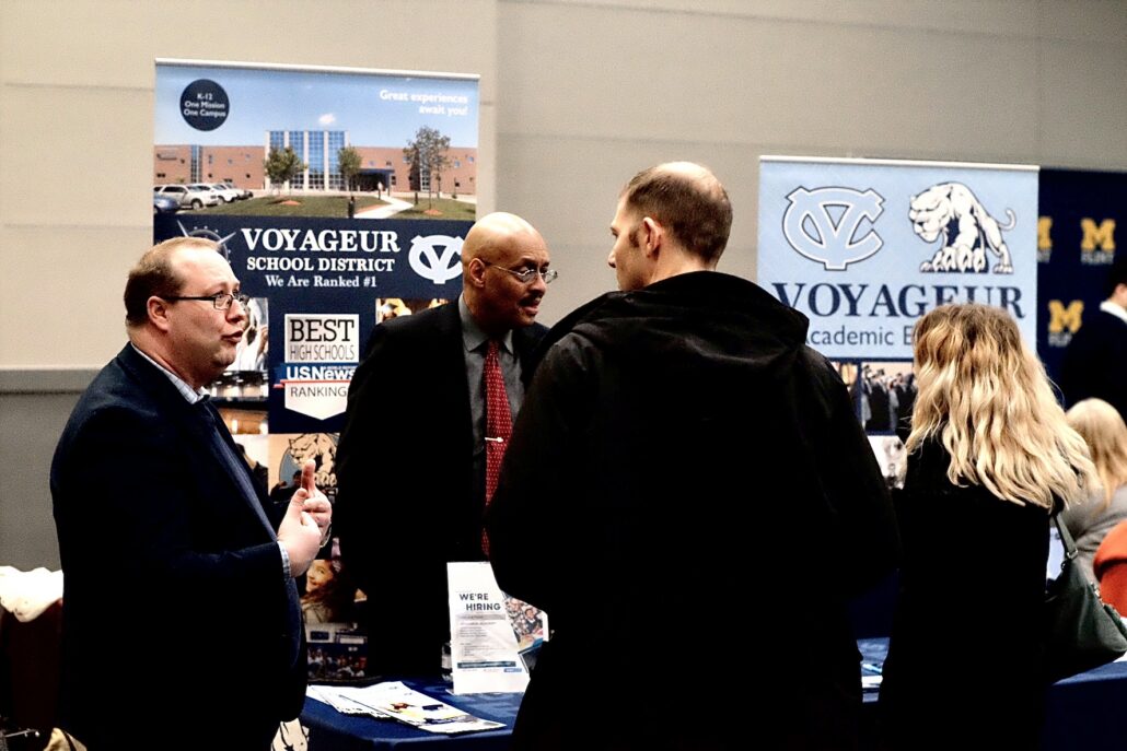 People talking at a booth for Voyageur School District with banners promoting the school and hiring opportunities.