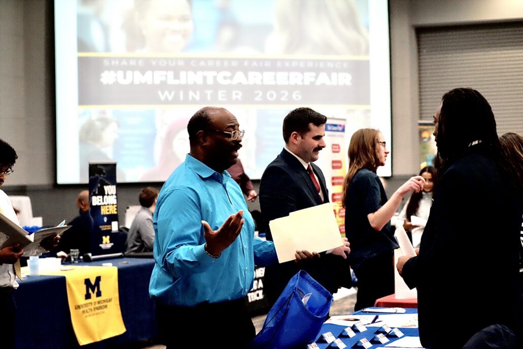 People interacting at a career fair with University of Michigan-Flint banners and a screen displaying "#UMFLINTCAREERFAIR Winter 2026" in the background.