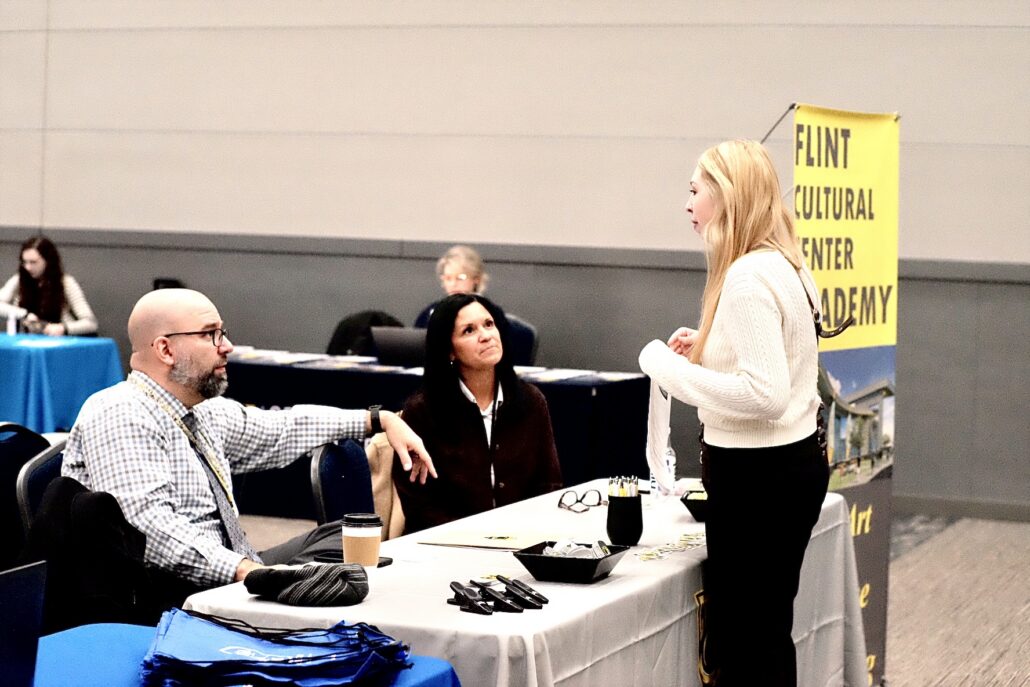 People interacting at a table with promotional materials and a yellow banner that reads "Flint Cultural Center Academy."