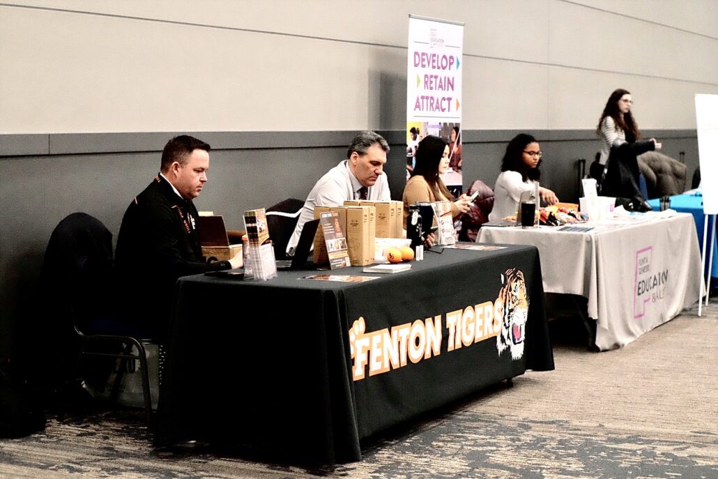 People sitting behind tables at an indoor event, one table has a black cloth with "Fenton Tigers" and a tiger logo, another table has a gray cloth with "Educate & Lead" text.