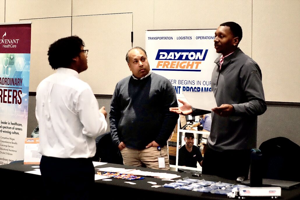 Three people standing and talking at a job fair booth with Dayton Freight and Covenant HealthCare banners in the background.