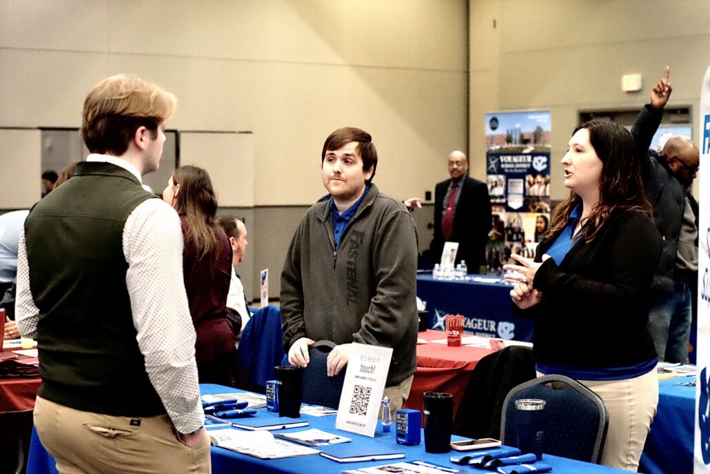People interacting at a career or information fair with tables displaying promotional materials and signs.