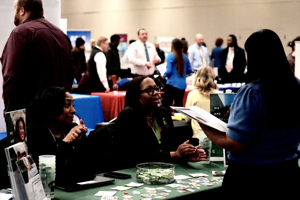 People interacting at a job fair or networking event with tables displaying promotional materials and brochures.