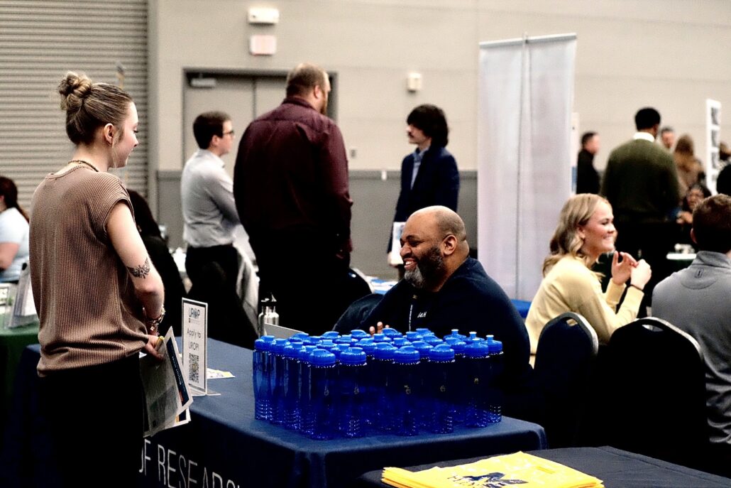 People interacting at a booth with a table covered in blue water bottles at an indoor event.