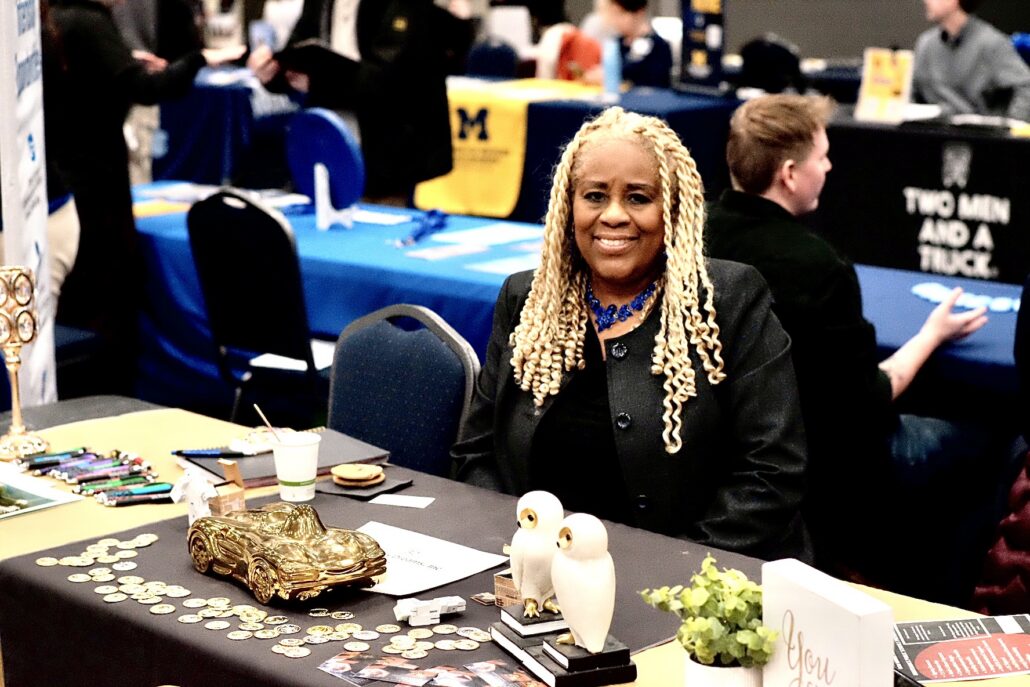 Person with long blonde braids sitting behind a table with a golden car model, two white owl figurines, coins, and a small plant at an indoor event.