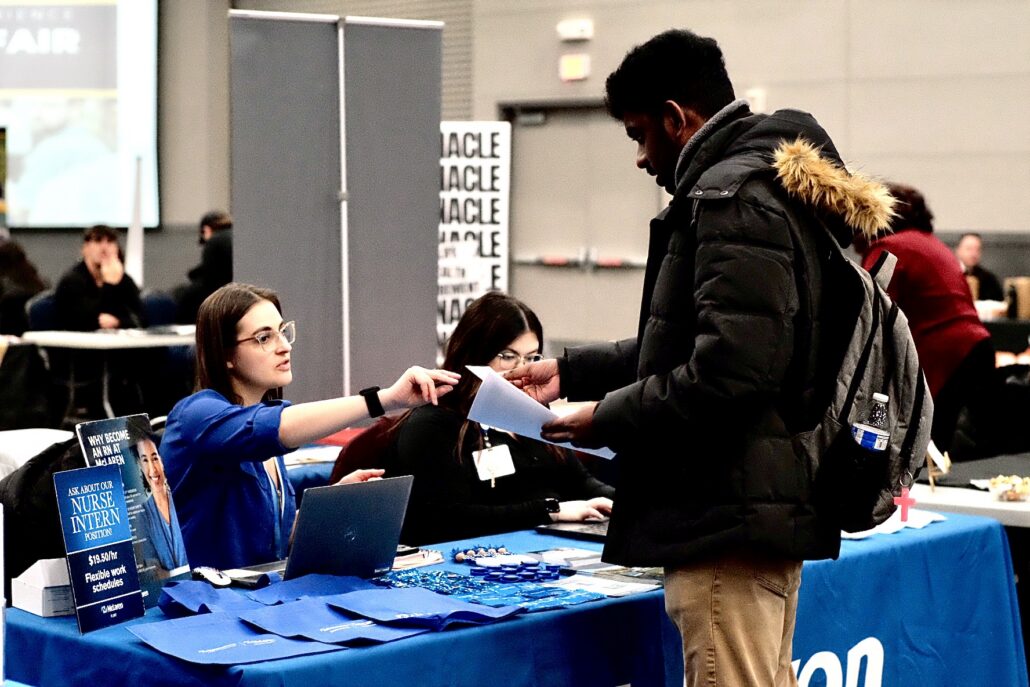 Person in a black jacket and backpack receiving a paper from a woman in a blue shirt at a nurse intern recruitment booth with informational materials on a blue tablecloth.