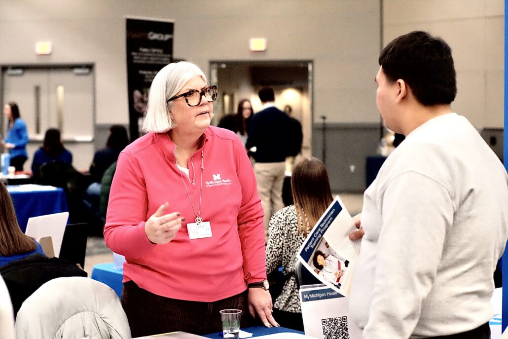 A woman in a pink MyMichigan Health pullover talks to a man holding a MyMichigan Health brochure at an indoor event.