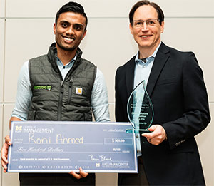 Two men holding a large check and a glass award, standing indoors against a light-colored wall.