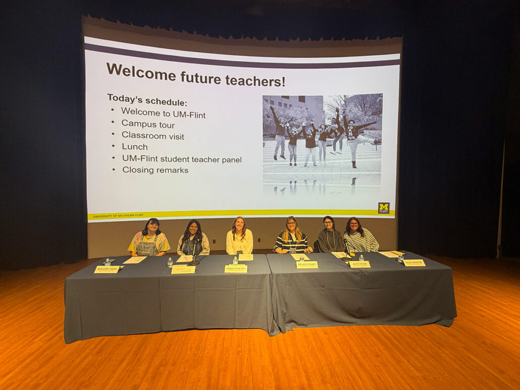 Panel of six people seated at a table on stage with a large screen behind them displaying a welcome message and schedule for future teachers at University of Michigan-Flint.