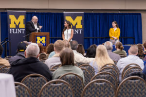 Audience watching a speaker at a podium with two women standing on stage in front of University of Michigan-Flint banners.