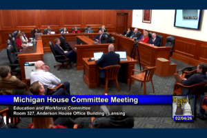 A Michigan House Committee meeting in session with members seated around a large wooden U-shaped table, a man speaking at a central desk with two laptops, and an audience seated in chairs. The screen shows the meeting is for the Education and Workforce Committee in Room 327, Anderson House Office Building, Lansing, dated 02/04/26 at 11:01 AM.