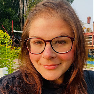 A person with long brown hair wearing glasses and a black top outdoors with greenery and a wooden structure in the background.