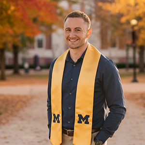 Person wearing a dark shirt and a yellow graduation stole with the letter M, standing outdoors with autumn trees in the background.