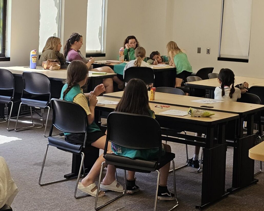 Classroom with children sitting at tables, some wearing green vests, engaged in activities and conversation.