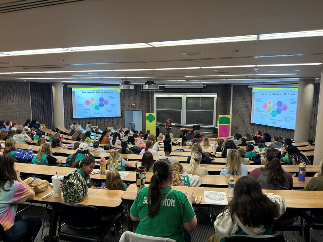 Lecture hall filled with students attending a presentation on "The Seven Pillars of Wellness" displayed on two large screens.