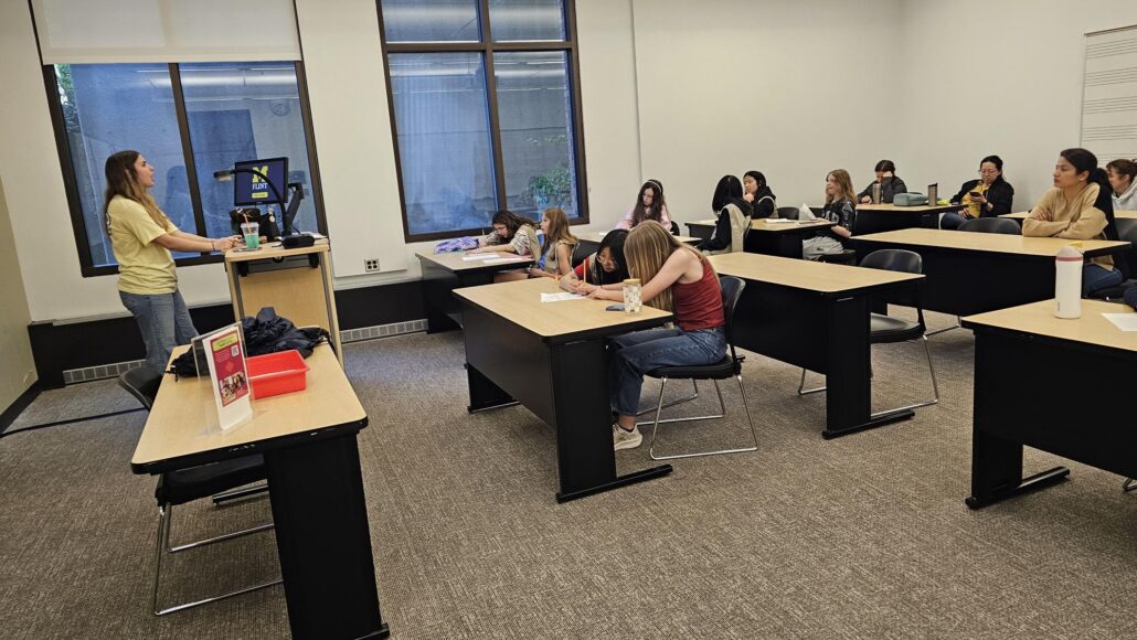 Classroom with students sitting at desks, some writing, and a person standing at a podium near a screen displaying the University of Michigan-Flint logo.