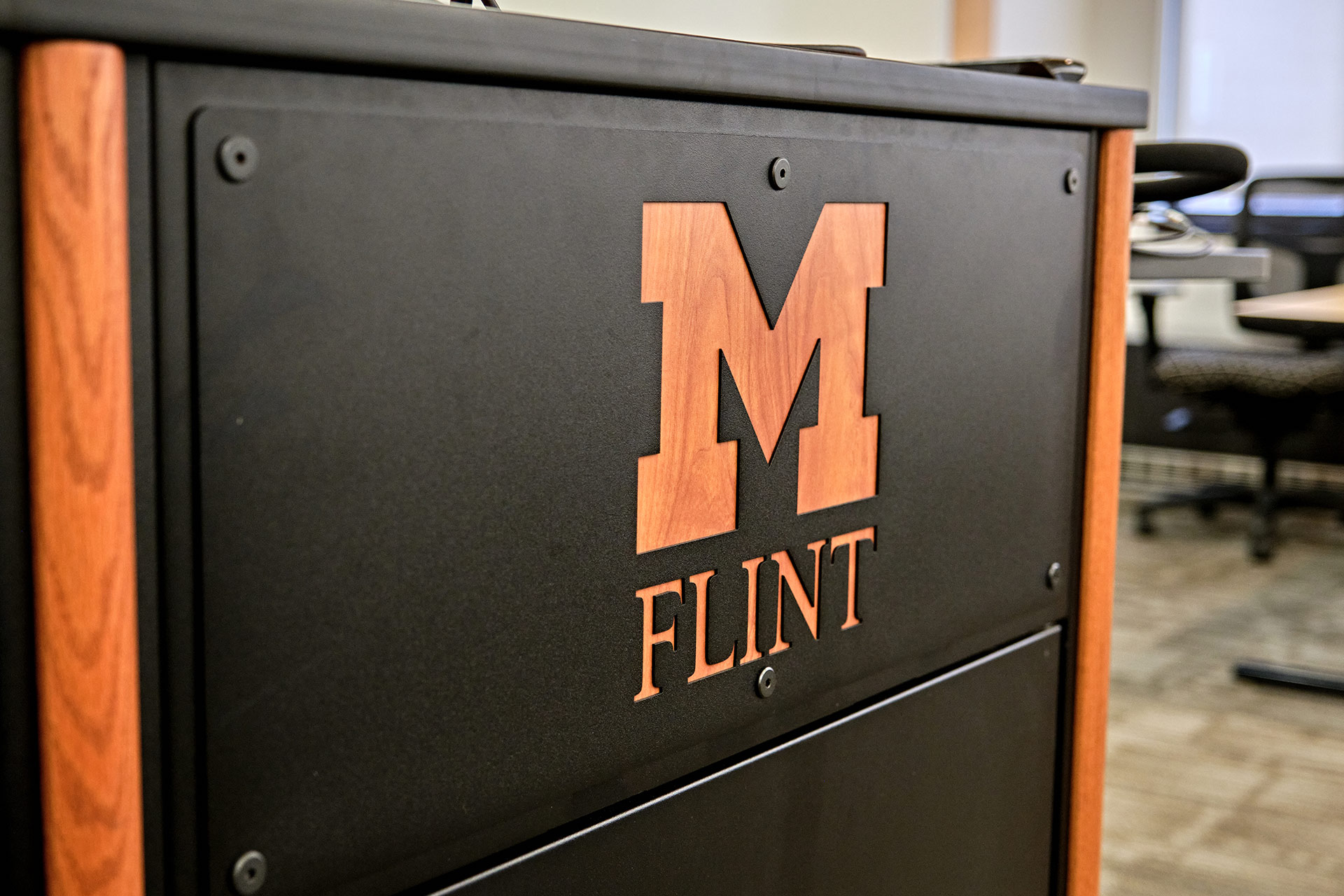 University of Michigan-Flint logo on a black and wood podium in an office setting.