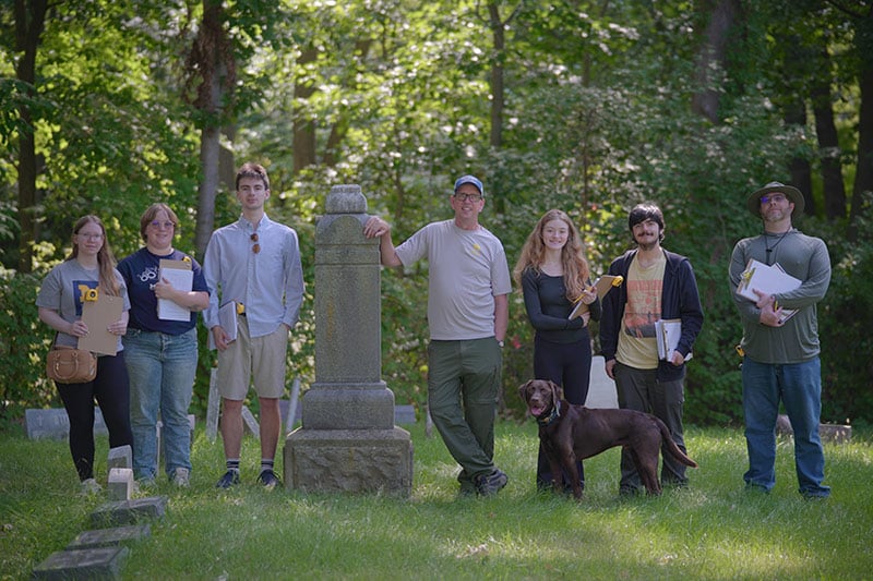 A group of seven people stands in a grassy area near a stone monument, with a dog in front of them, surrounded by trees.