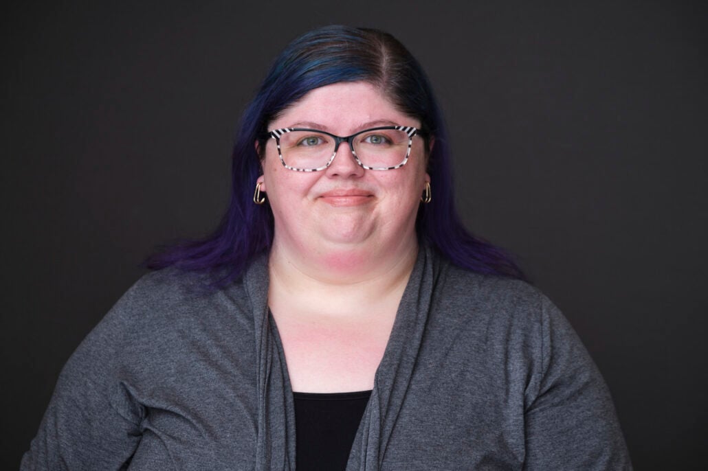 A person with glasses and purple hair smiles against a dark background, wearing a gray cardigan over a black top.