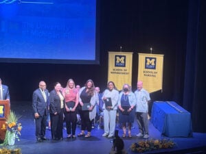A group of six people stands on stage at a ceremony, holding awards, with banners for the University of Michigan-Flint in the background.