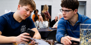 Two people are sitting at a table, one looking at a phone while the other observes, with a group of people in the background.