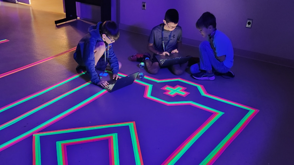 Three children are sitting on a floor with colorful neon lines, using laptops in a dimly lit room.