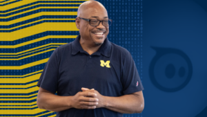 A person smiles while standing with hands clasped, wearing a dark shirt with a yellow "M" logo, against a tech-themed background.