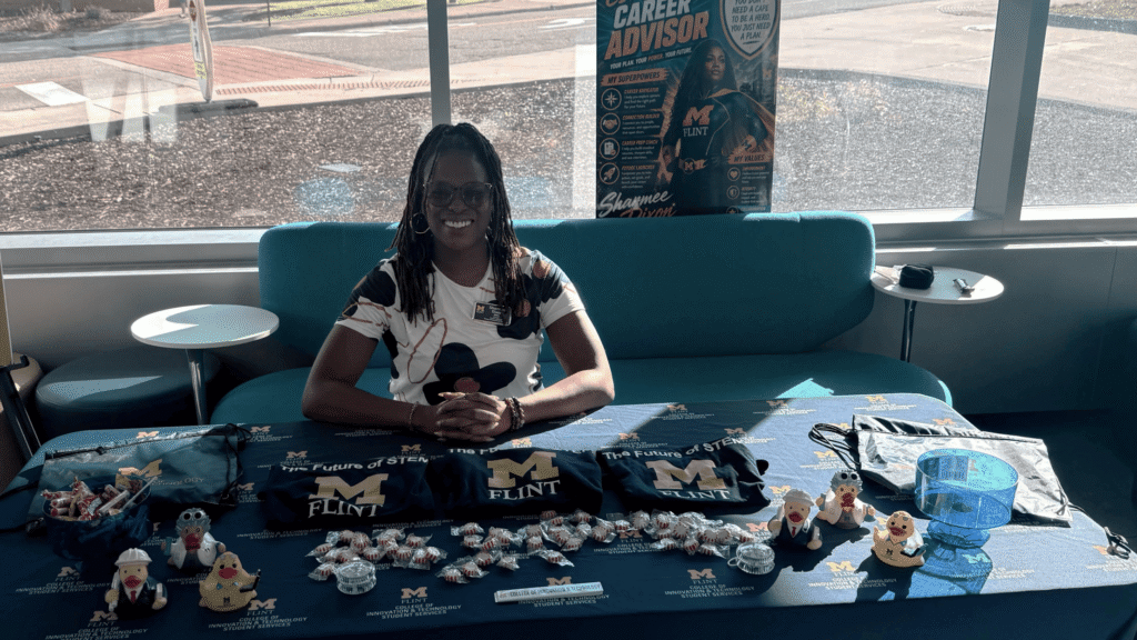 A person sits at a table displaying promotional items and materials for the University of Michigan-Flint, with a poster in the background.