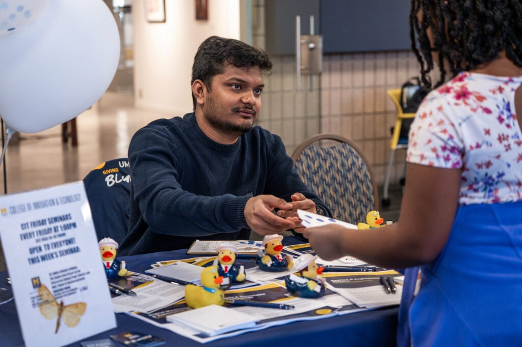 A person hands a card to another person at a table with promotional materials and rubber ducks, in a bright indoor setting.