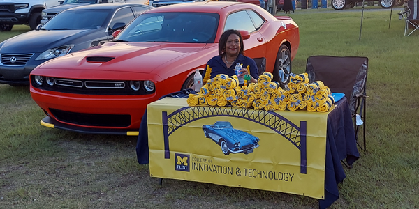 Shirl Donaldson sits behind a UM-Flint College of Innovation & Technology table loaded with T-shirts with a red Dodge Challenger GT behind her.