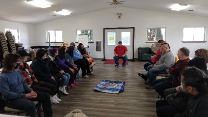 Two rows of people sit face-to-face about 8 feet apart with a quilt of sorts in the middle of the floor between them. A man sits between the rows at the far end of the room and speaks to the group of 12-15.