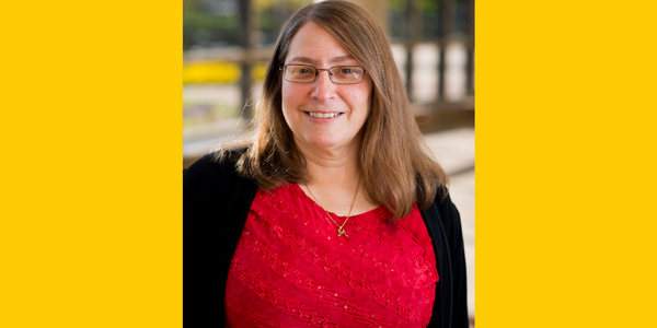 yellow background, faculty headshot of Michelle Sahli smiling in a red shirt with a black blazer in the Atrium of the library.