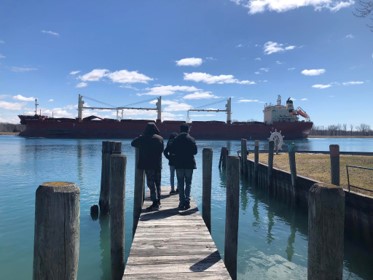 A few students stand on a dock and look off in the distance at a big cargo ship.