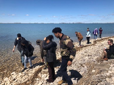 Students look down at the rocks beneath them with a bunch of water past them and the Detroit skyline past that.