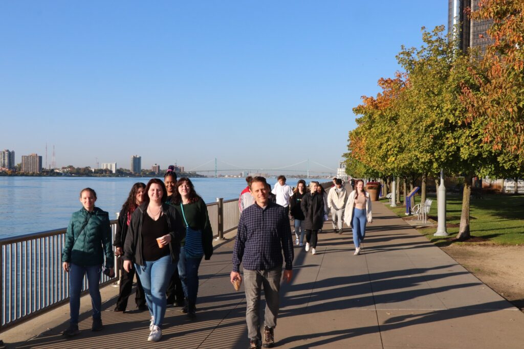 Lederman leads students on the 'River walk' with the Rennaisance building on the right behind fall trees and the city of Windsor across the water to the left.