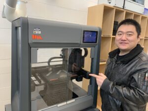 UROP research assistant Joon Soo Park points to the 3D printer in process and smiles. It has a class encased box with a raised tray and the printer is on a track above the tray.
