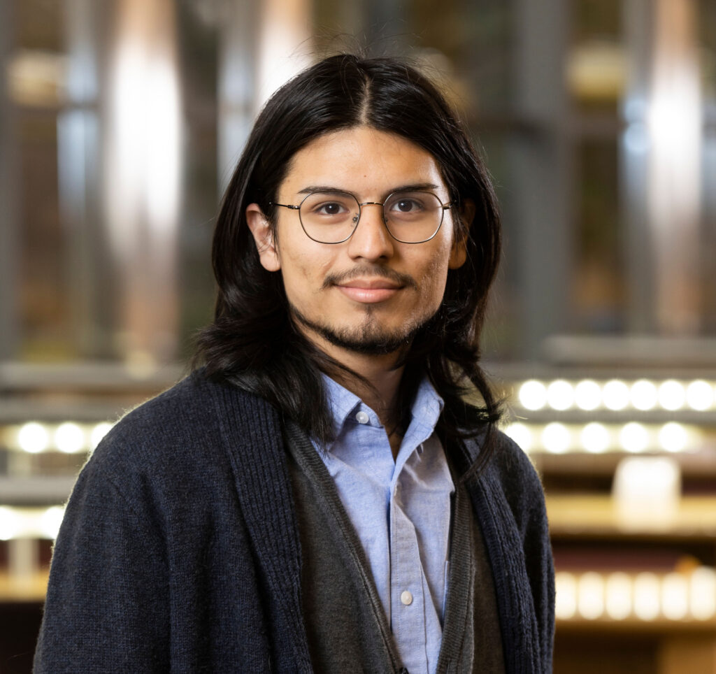 Luis Martinez poses in the Library atrium for a headshot photo. They have shoulder-length black hair parted in the center, wire-rimmed glasses, a thin moustache and chin-strap beard. They are wearing a light-blue collared shirt with a grey vest and dark-blue sweater over that.