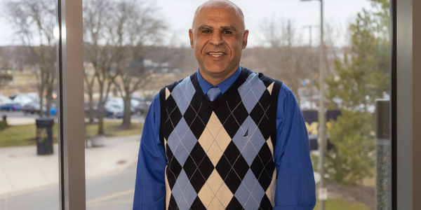 Khalil Khanafer poses in the MSB for a headshot. He is smiling and wearing a blue shirt with a blue, creme, and argyle vest on.