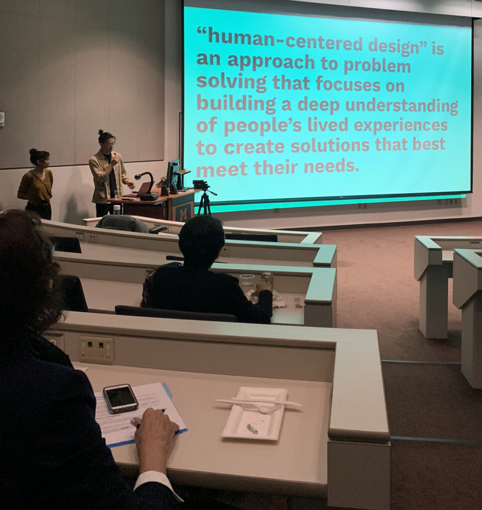Viewers look on as Nakagawa and Uroda present in a large conference room. They stand to the left of a large projection screen, which says, "human-centered design" is an approach to problem solving that focuses on building a deep understanding of people's lived experiences to create solutions that best meet their needs."