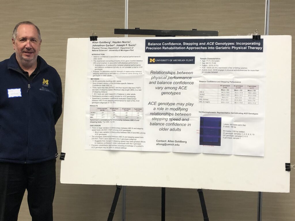 Man stands in blue pullover fleece with M-Flint logo on it. His arms are clasped behind his back. He's smiling. To his right there is a research poster.