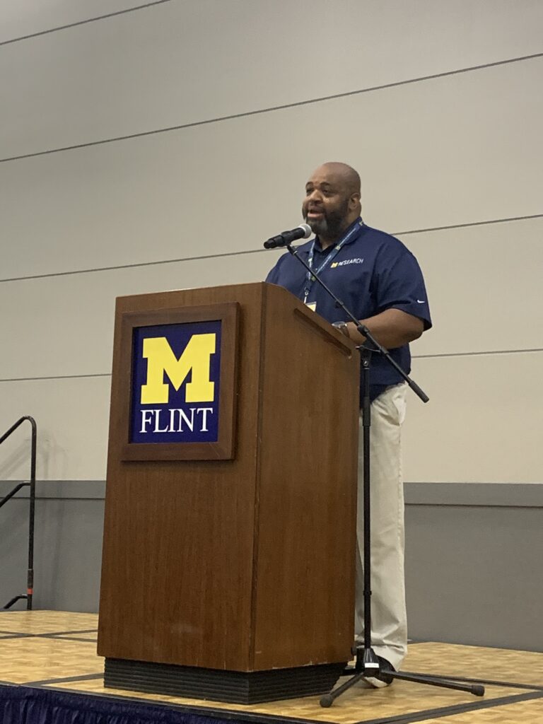 A man stands behind a tall podium with the UM-Flint block logo on it. He is wearing a blue collared shirt, khaki pants, and a broad smile.